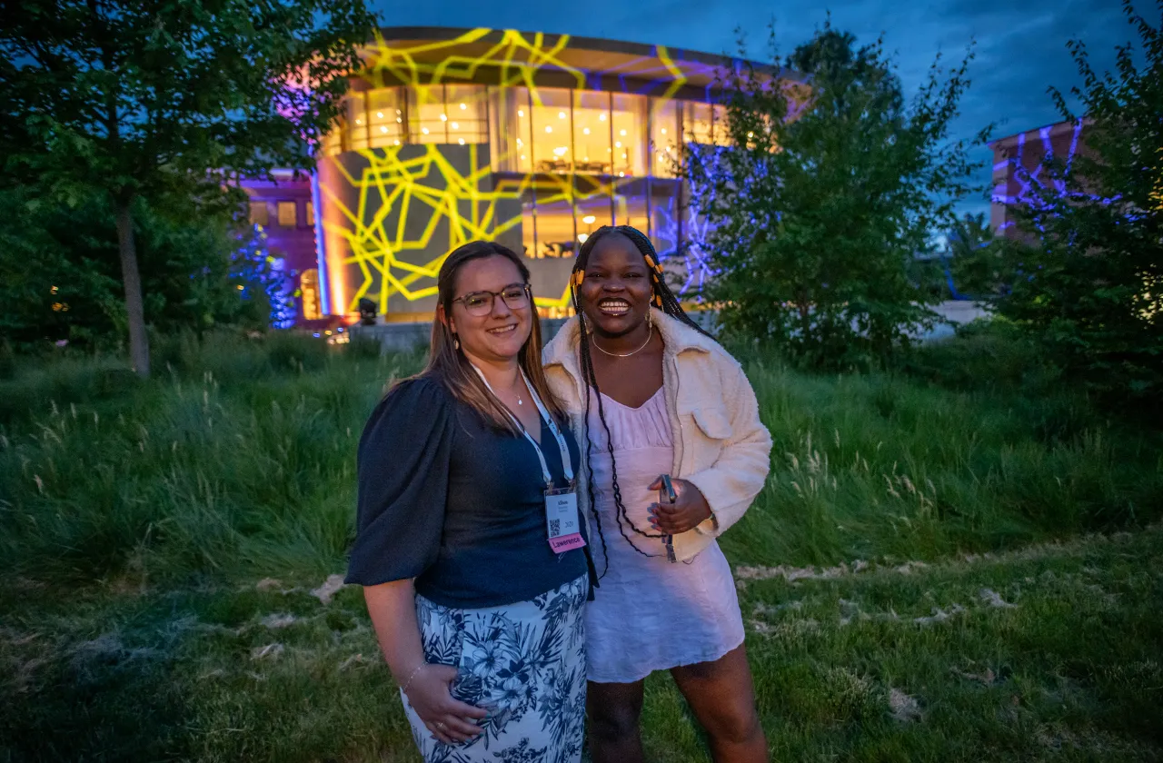 Two alums stand in front of Neilson Library, which is illuminated with colorful patterns.