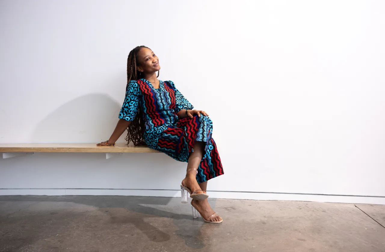 Adwoa Pokuaa Boakye sits on a bench against a white wall while wearing a red and blue printed outfit.