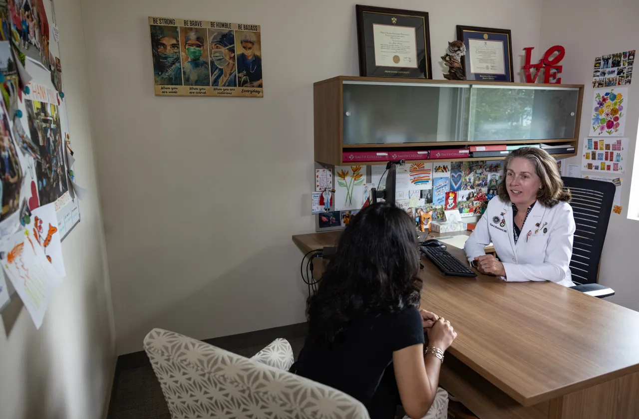 A doctor in a white lab coat sits behind a desk, talking with a student