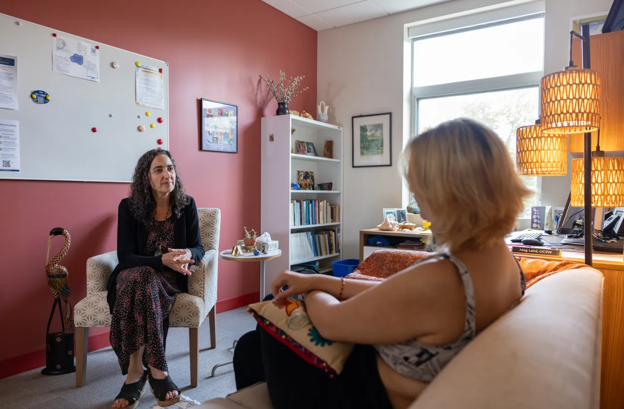 A counselor sits on a chair and talks with a client, who is holding a pillow in their lap