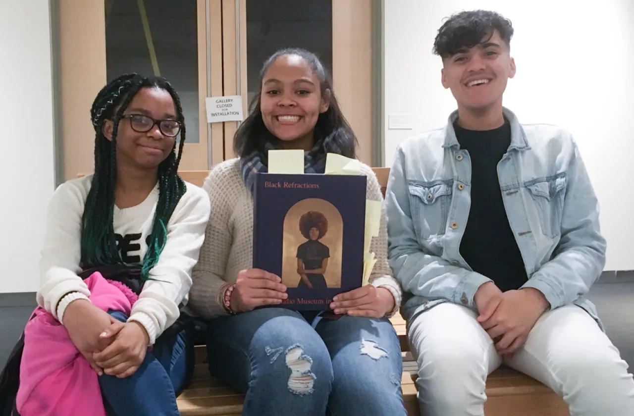 Three students of color seated and smiling at the camera. The middle person holds a copy of the exhibit book Black Refractions