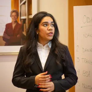A woman in a black suit speaks confidently by a whiteboard with handwritten names. A portrait of another woman is visible in the background.