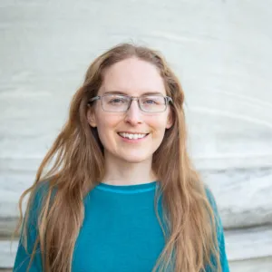 Portrait of a smiling woman with long strawberry blond hair, thin-framed glasses, and a teal shirt, standing in front of a light gray stone column.