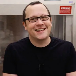 Portrait of a smiling man with short dark hair, black rectangular glasses, and a black t-shirt, standing in front of laboratory equipment with a red warning sign.