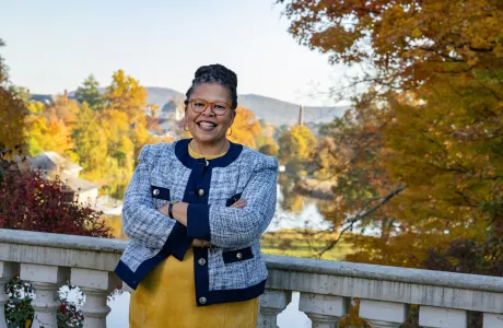 Sarah Willie-LeBreton smiles toward the camera in front of fall foliage, leaning on a railing and crossing her arms, wearing a yellow shirt with a blue jacket on top.