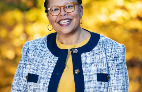 Sarah Willie-LeBreton smiles toward the camera in front of fall foliage, wearing a yellow shirt with a blue jacket on top.