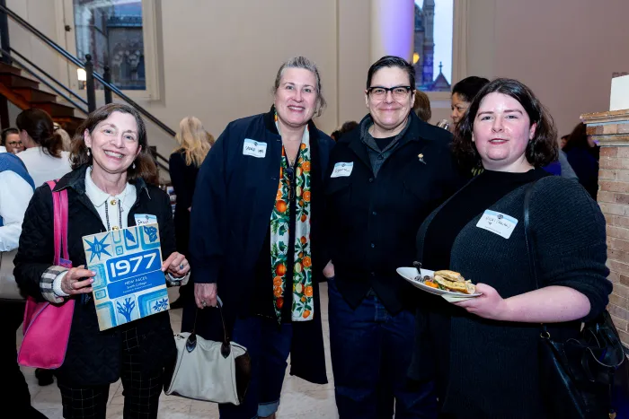 Four alums smile at the camera. One holds a sign that says "1977."