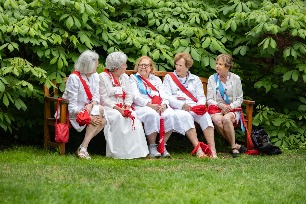 Smithies in white wearing red sashes sit and chat on a bench