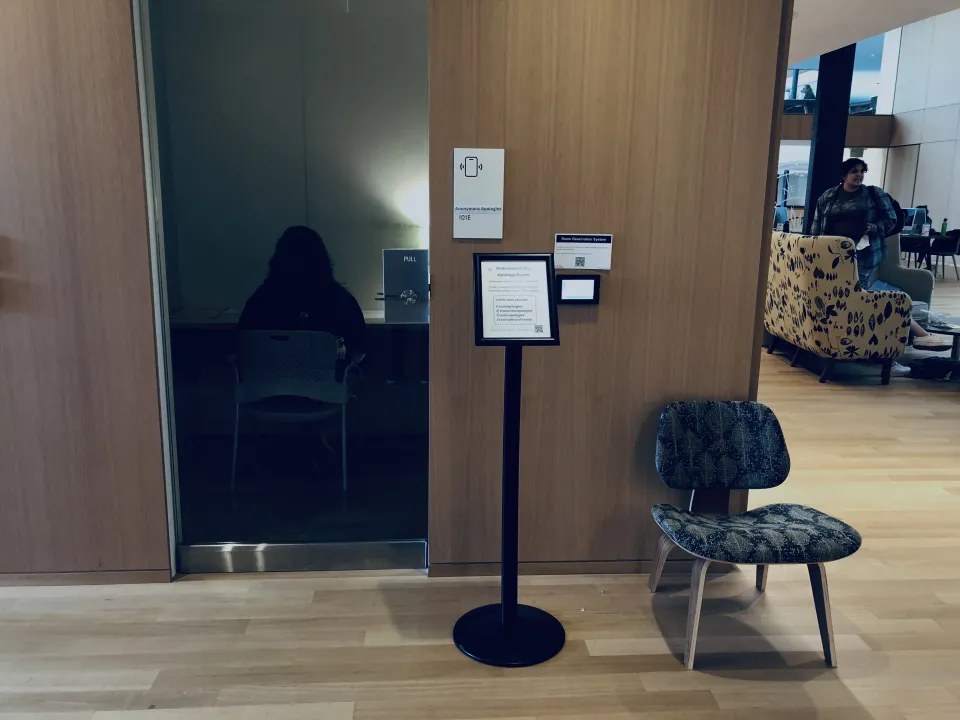 A chair, a small message on a black stand and a shadowy figure in the Anonymous Apologies space in Neilson Library