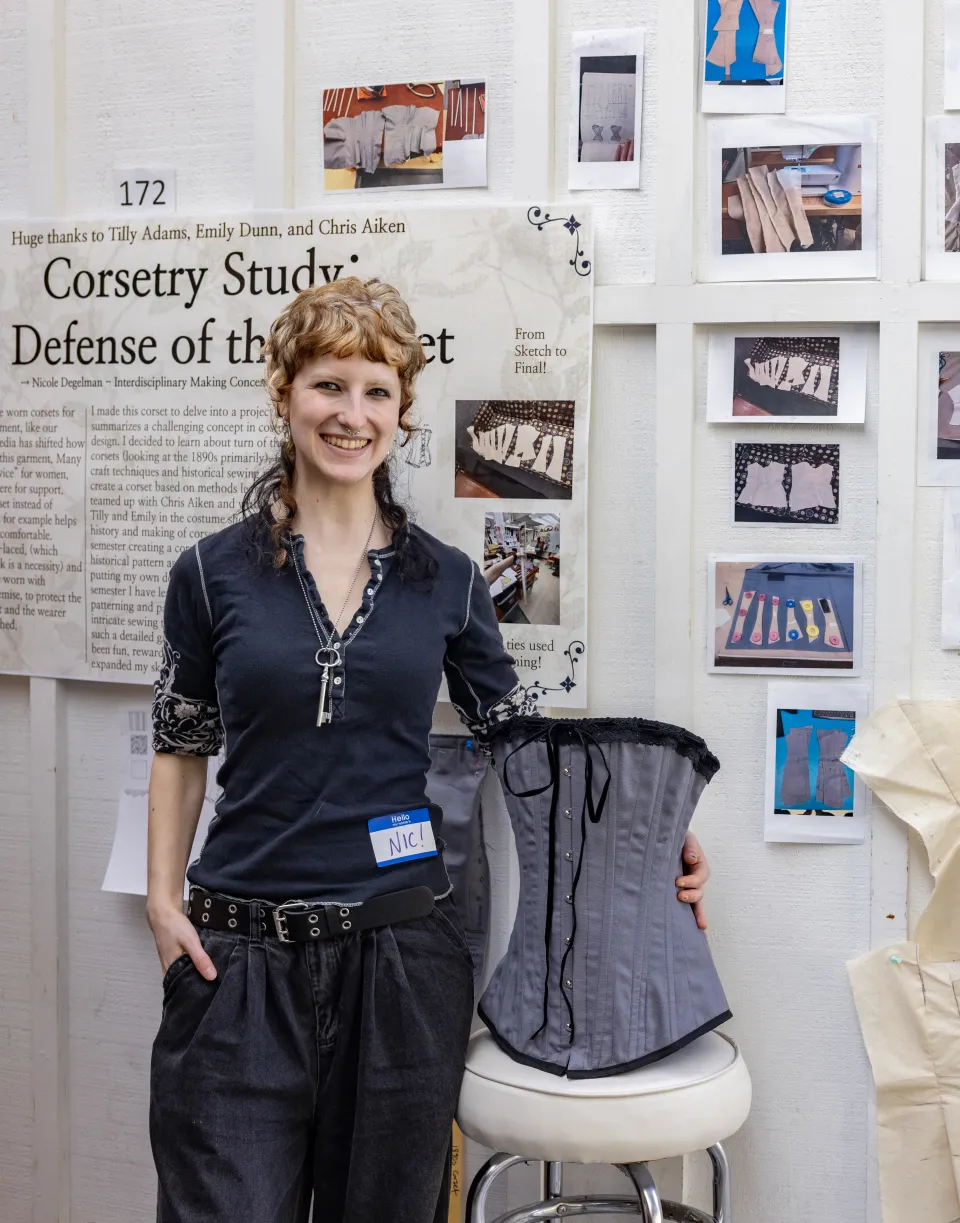 Nic Degelman stands in front of her presentation for Collaborations, holding a handmade corset