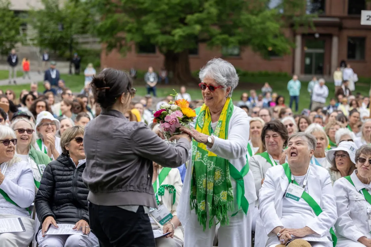 A woman with sunglasses and a green scarf receives a bouquet, smiling warmly. She's surrounded by a seated crowd, creating a joyful atmosphere.