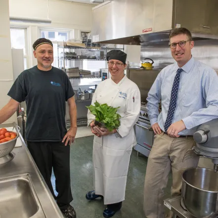 Three members of Dining staff in a kitchen. One man is near a bowl of tomatoes, and a woman is holding a bundle of greens.