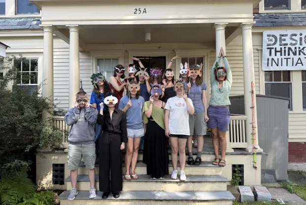 A group of about thirteen people pose on the porch steps of a light yellow building with columns labeled "25A". Every person is holding or wearing a handcrafted paper animal or decorative mask over their face. A banner on the right side of the building reads THE DESIGN THINKING INITIATIVE.